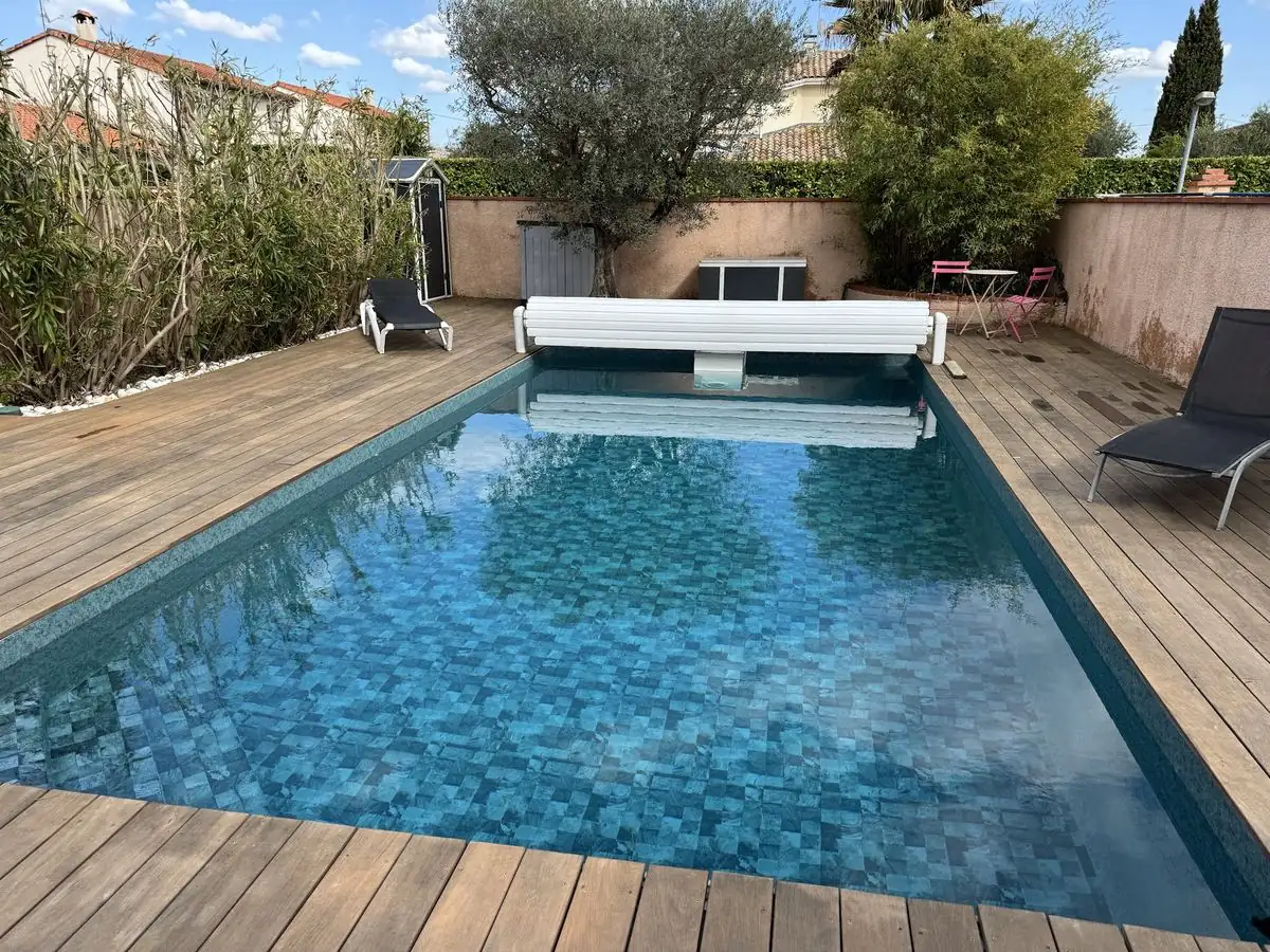 Piscine terminée avec revêtement mosaïque bleu et plage en bois dans un jardin.